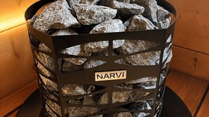 Een zwarte metalen mand met het label 'NARVI', gevuld met stenen, geplaatst op een ronde voet tegen een houten wand.