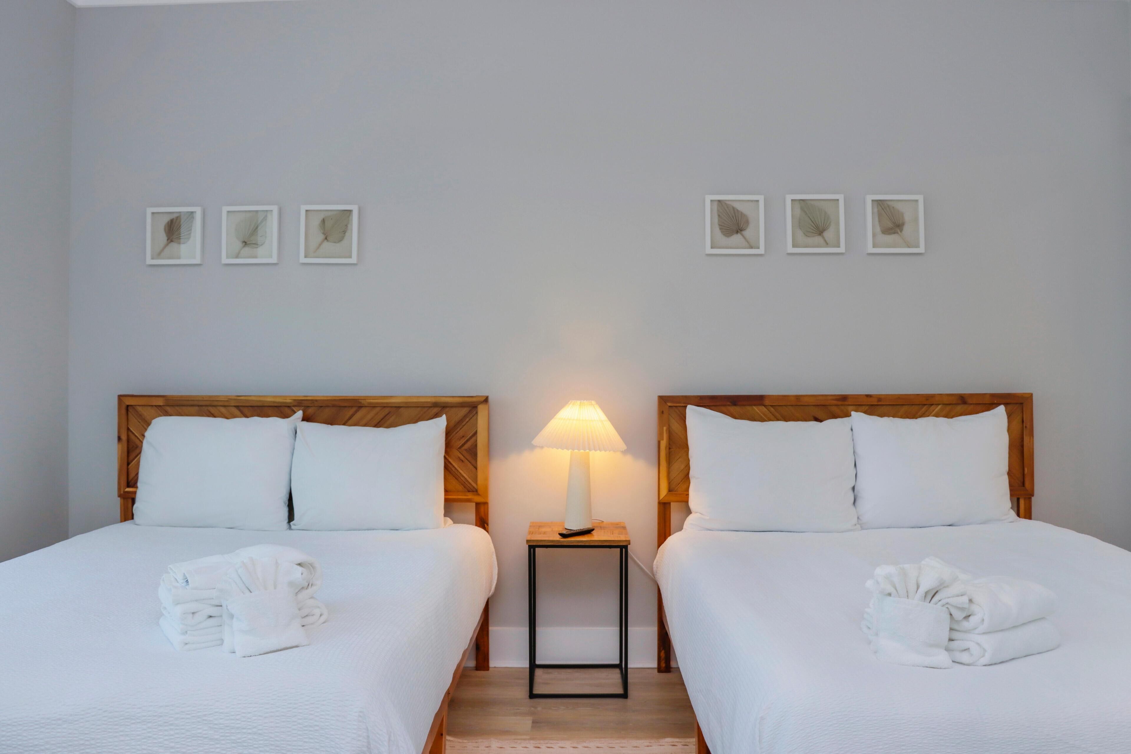 Habitación de hotel con dos camas, una mesita de noche con lámpara y cuatro cuadros enmarcados en la pared.
