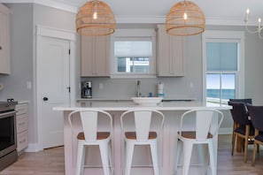 A modern kitchen with white cabinetry, a large island with bar stools, and a view of the ocean through a large window. - Windward Crest (Port St. Joe)