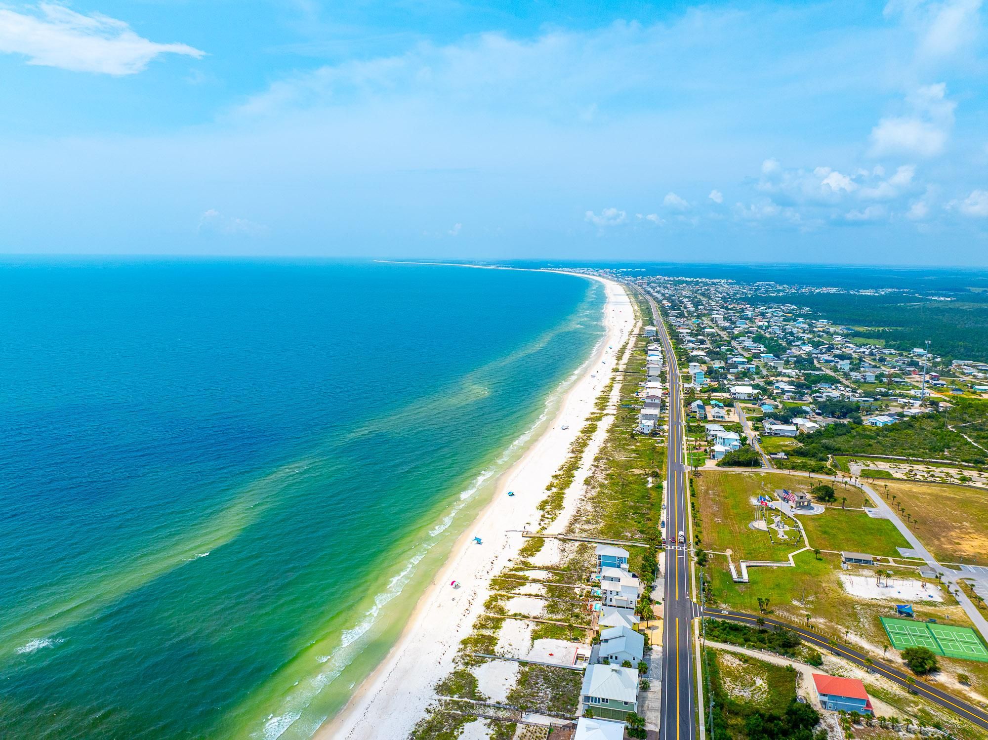 Eine Luftaufnahme einer Küstenregion mit einem langen Strand, einer parallel zur Küste verlaufenden Straße und einem dicht bebauten Wohngebiet mit Häusern und viel Grün.