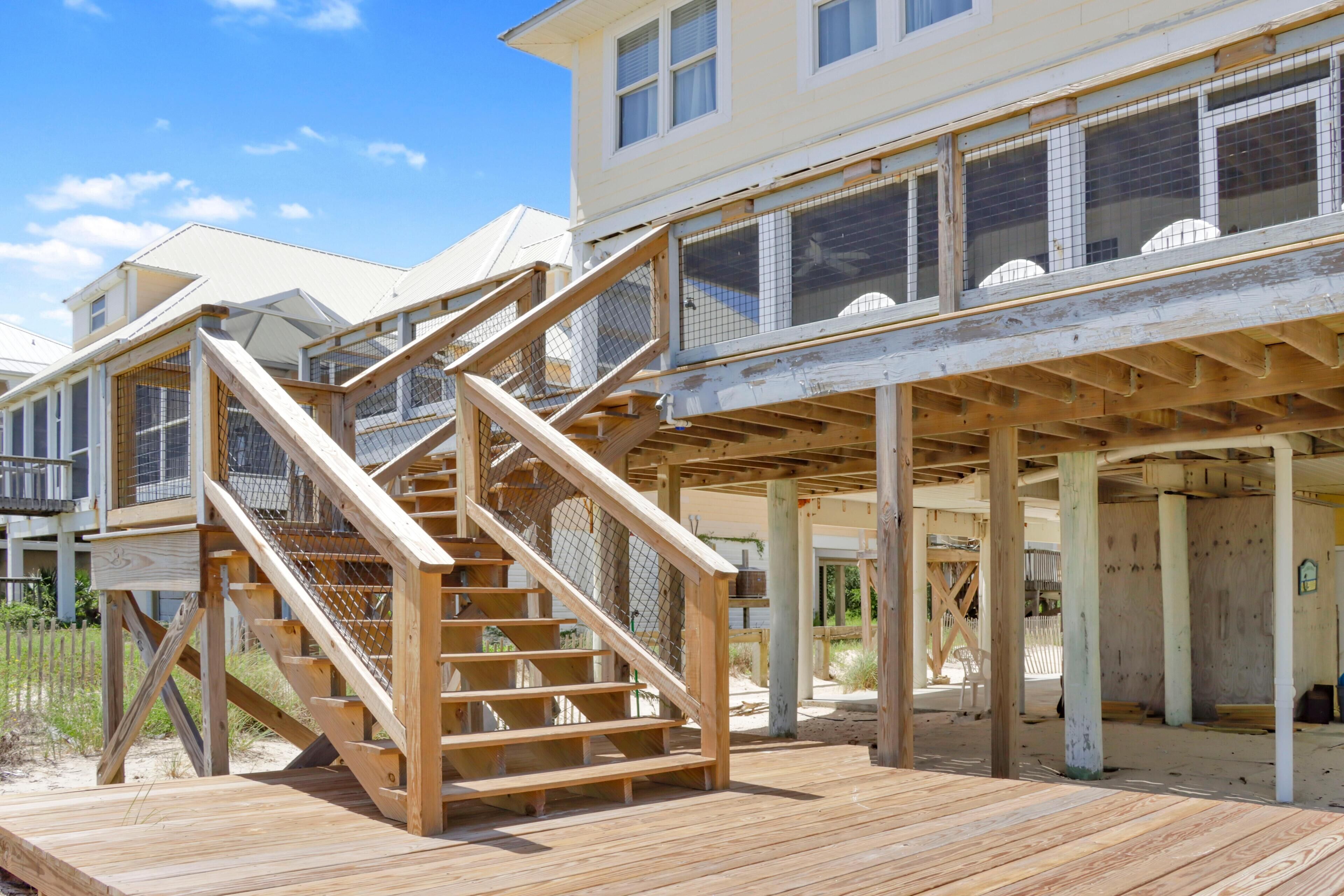 Eine Holztreppe führt zu einer überdachten Terrasse mit Blick auf einen Strand und ein Gebäude mit mehreren Fenstern.