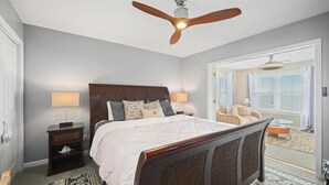 Un dormitorio con una cama grande, dos lámparas de noche, un ventilador de techo y vistas al salón contiguo.