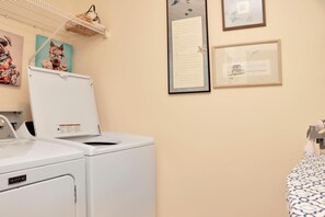 Ein Waschbereich mit einer weißen Waschmaschine, einem Regal mit einem Wäschekorb und einem Bett mit einem gemusterten Bettbezug.