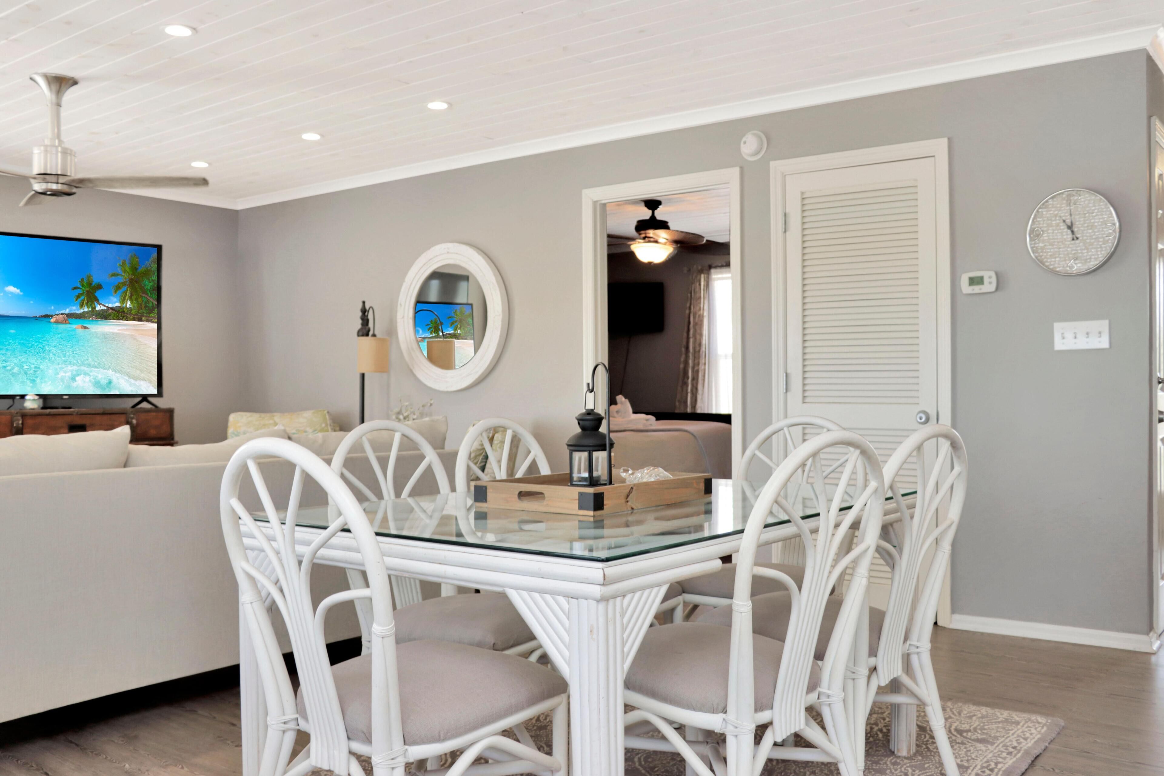 Un comedor con mesa de vidrio y sillas de mimbre blanco, un televisor mostrando una escena de playa, y una puerta que da a otra habitación.