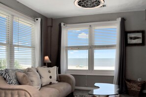 Ein Zimmer mit einer Couch, einer Lampe, einem Fenster mit Blick auf den Strand und einem Deckenventilator.