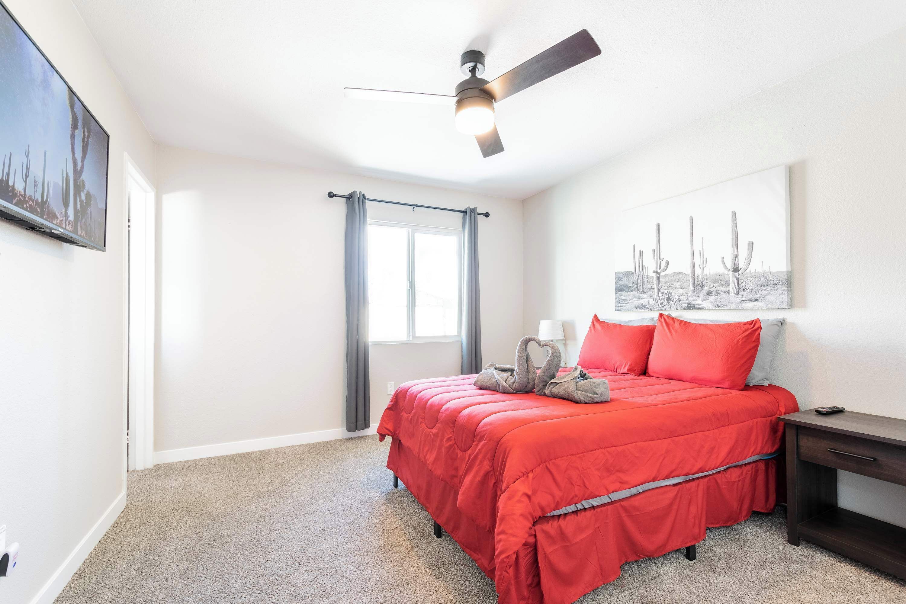 A bedroom with a bed, a ceiling fan, a window with curtains, a nightstand, and a wall-mounted TV.
