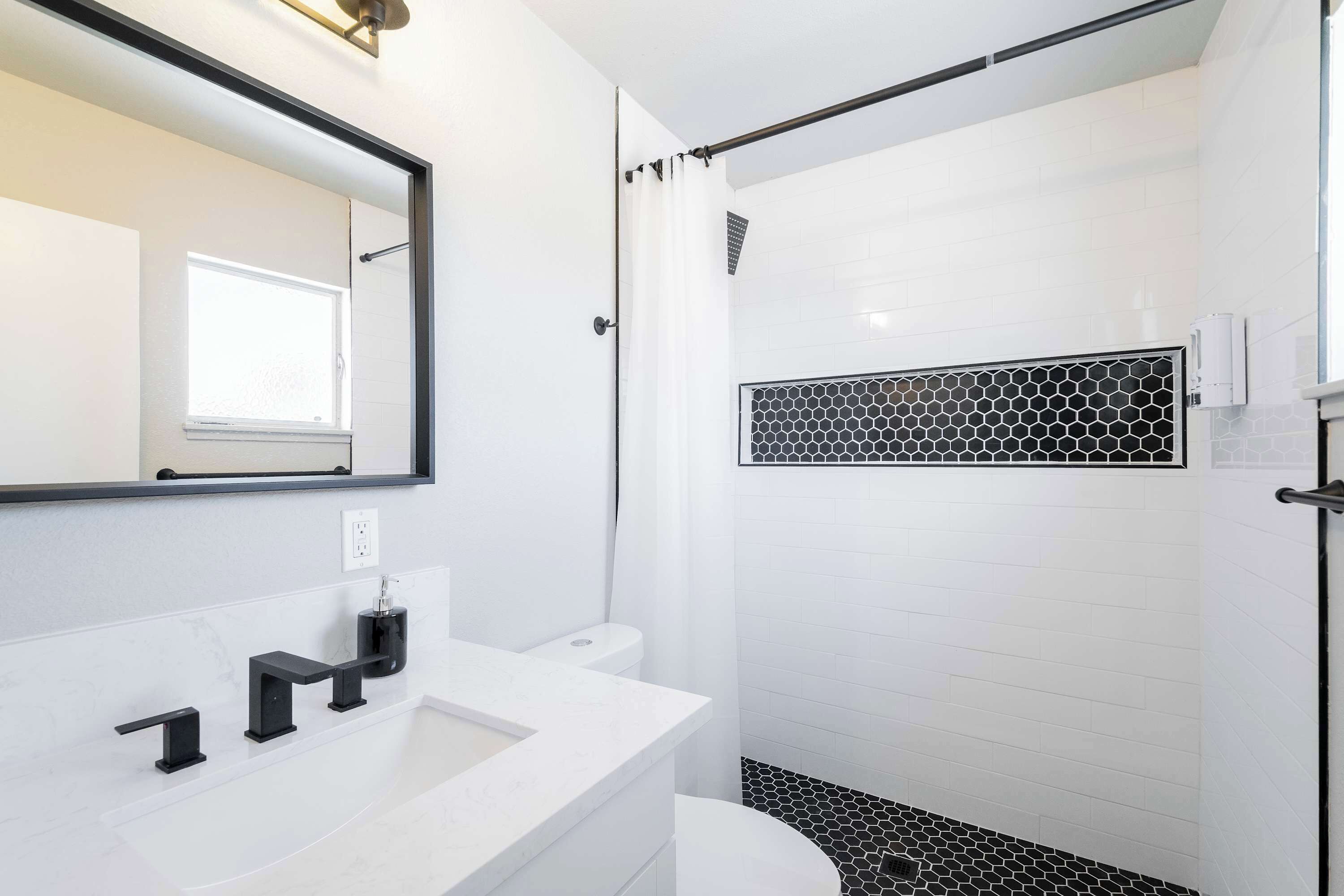 Un baño moderno con lavamanos blanco, accesorios negros, ducha con azulejos hexagonales y un espejo grande.