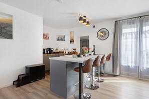 A modern kitchen with a central island, bar stools, a clock, and artwork on the walls. - Stay at "L'Ensoleillé" – City Center / Train Station (Les Sables-d'Olonne)