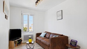 Een woonkamer met een bruine bank, een televisie op een houten kast, een klein rond tafeltje met een gele vaas en een raam met uitzicht op gebouwen.