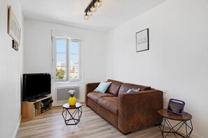 A living room with a brown sofa, a television on a wooden stand, a small round table with a yellow vase, and a window with a view of buildings. - Stay at "L'Ensoleillé" – City Center / Train Station (Les Sables-d'Olonne)