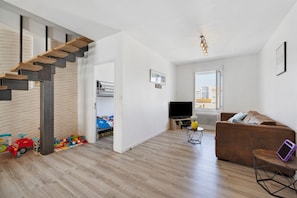 A modern living room with a wooden staircase, a sofa, a TV, and a bookshelf. - Stay at "L'Ensoleillé" – City Center / Train Station (Les Sables-d'Olonne)