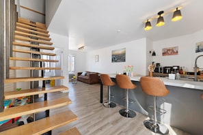 A modern kitchen with bar stools, a wooden staircase, and a view of the cityscape. - Stay at "L'Ensoleillé" – City Center / Train Station (Les Sables-d'Olonne)