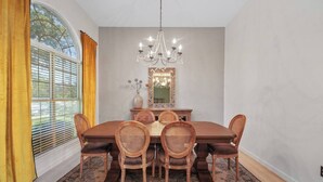 Uma sala de jantar com uma mesa grande, seis cadeiras, um candelabro, um espelho e uma janela com persianas.