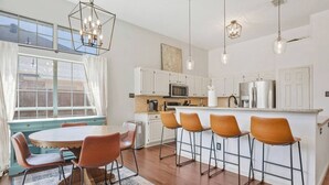 Uma cozinha moderna com armários brancos, uma ilha central com banquetas, uma mesa de jantar com cadeiras e luminárias pendentes.