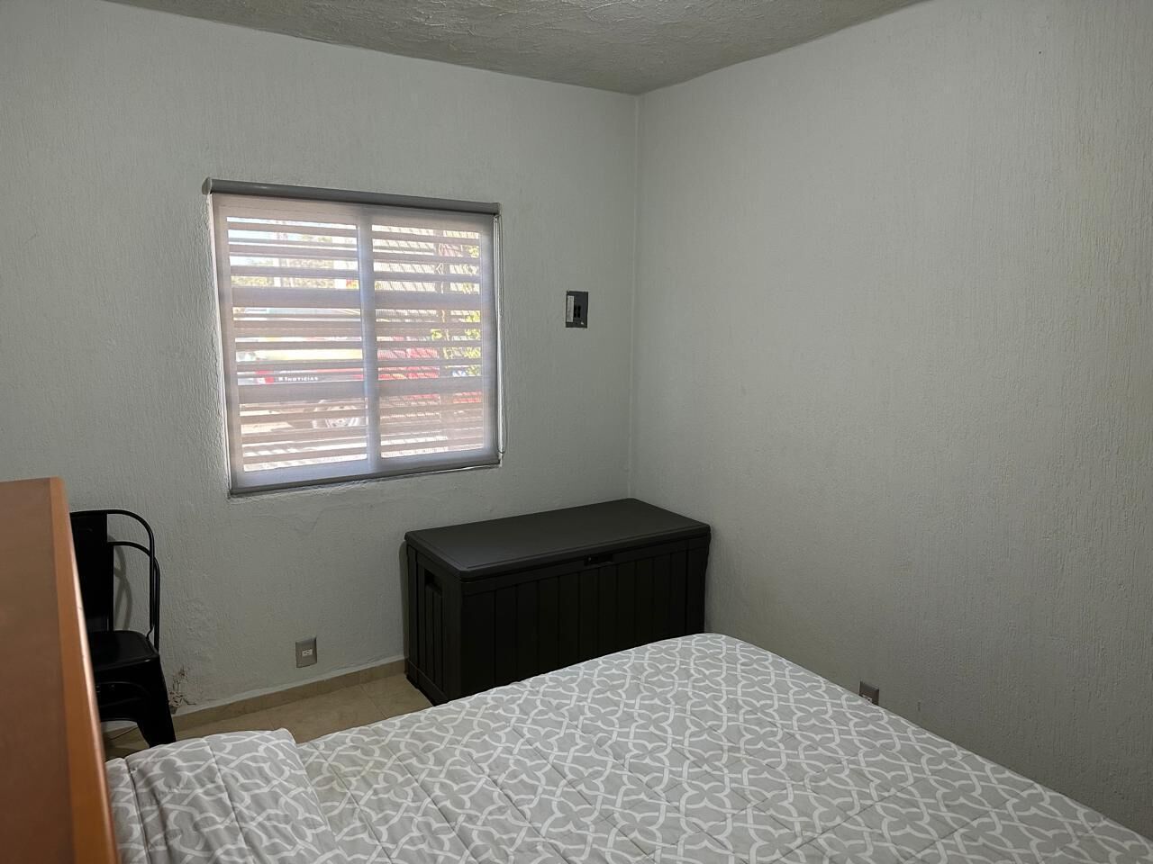 A bedroom with a bed, a window with blinds, a black dresser, and a chair.