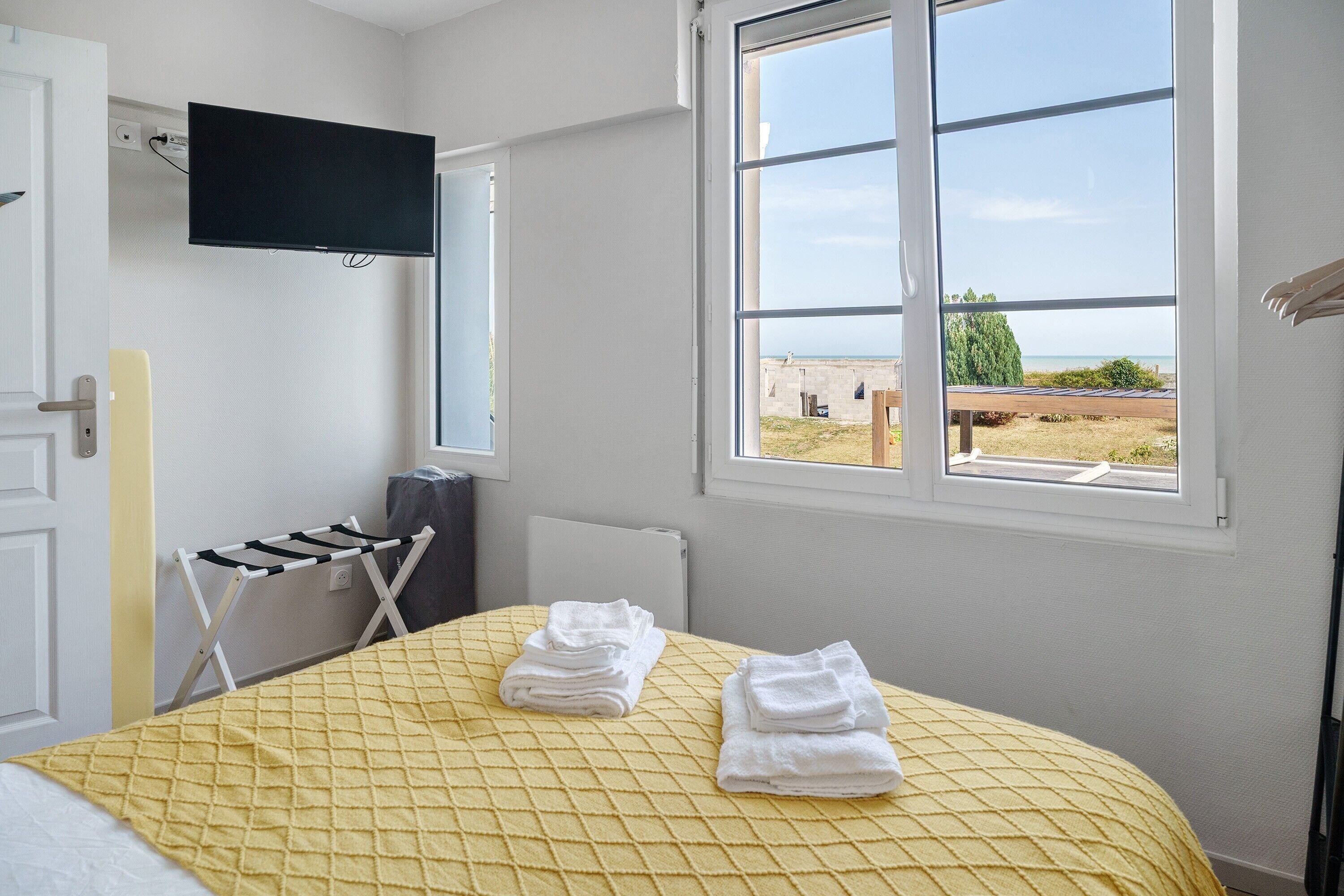 A hotel room with a bed, a TV mounted on the wall, a window with a view of the sea, and a drying rack.