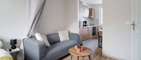 Een moderne woonkamer met een grijze bank, een klein rond houten tafeltje en een keuken op de achtergrond.