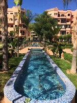 Una piscina con borde de azulejos, rodeada de palmeras y un edificio con balcones.