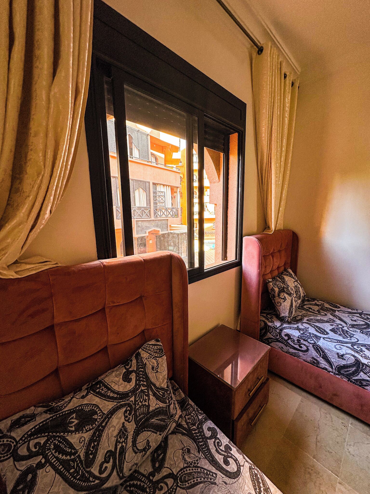 A room with two beds, a window, and a view of a building outside.