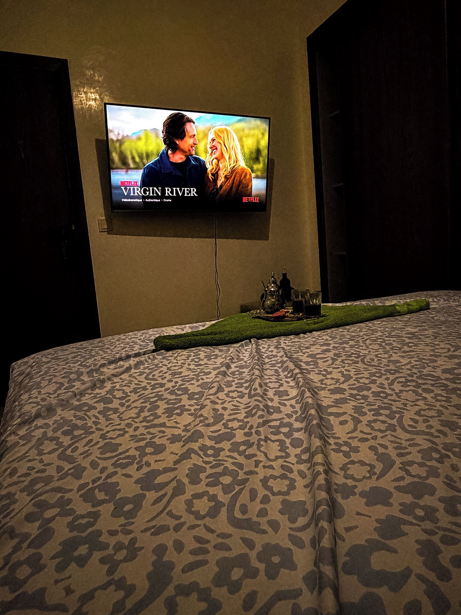 A television mounted on a wall displaying 'Virgin River' with a bed featuring a leopard print bedspread in the foreground.