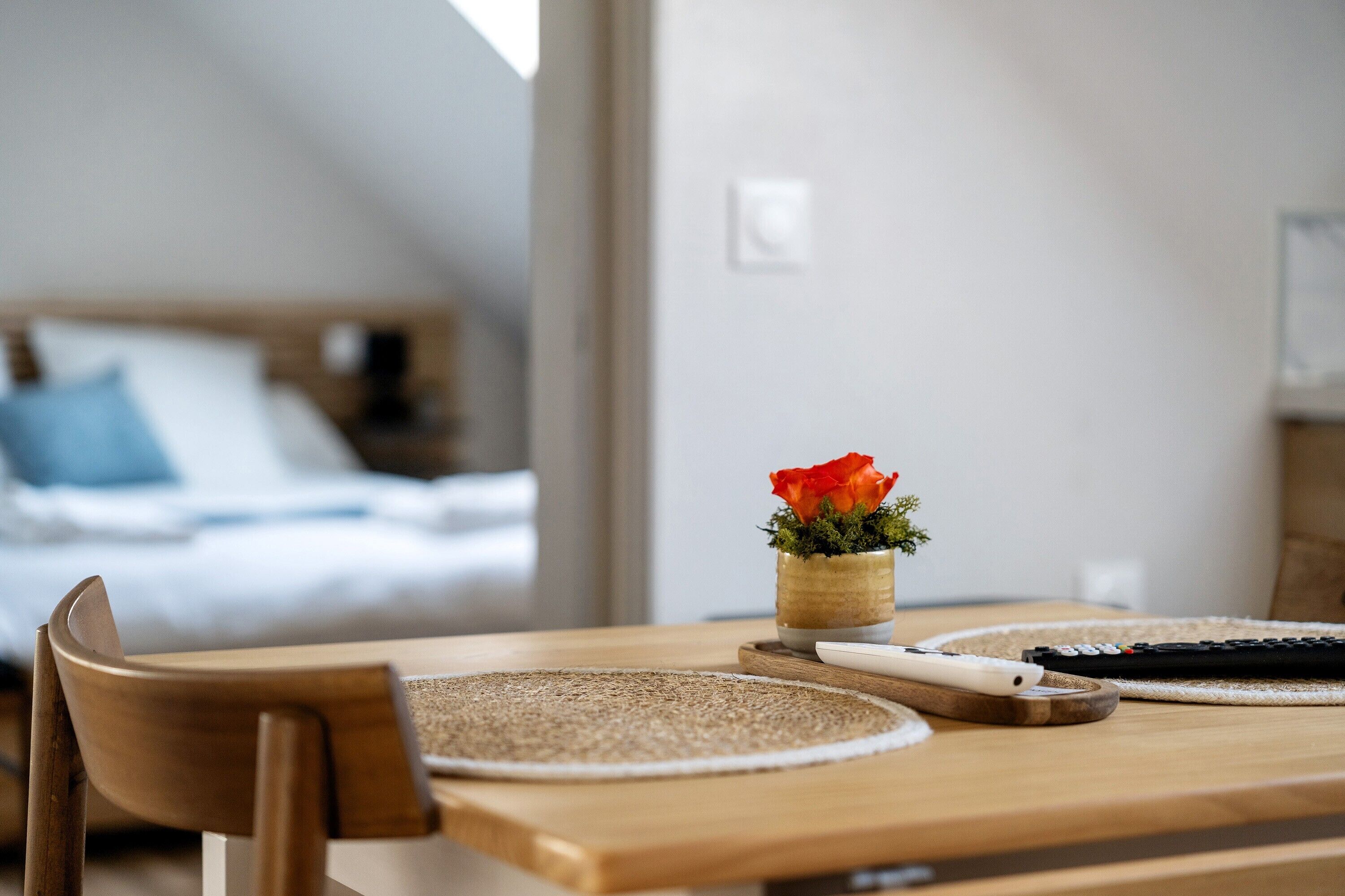 A wooden dining table with a vase of red flowers, a white plate, and a remote control.