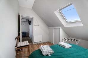 Ein Schlafzimmer mit schrägen Wänden, einem Oberlicht, einem Bett mit grüner Steppdecke, einem Holzstuhl und einem Wäscheständer.