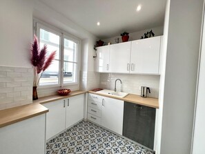 Eine kleine Küche mit weißen Schränken, einer Spüle, einer Kaffeemaschine und einem Fenster mit Zierpflanzen.
