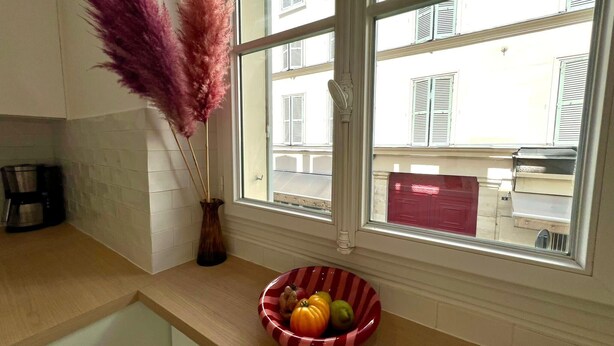 Ett kök med ett fönster, en vas med torkade växter och en skål med frukt på bänkskivan.