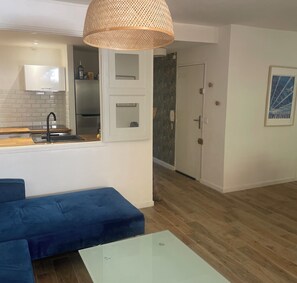 Ein moderner Wohnraum mit einer Küche, einem blauen Sofa, einem Glastisch und einer Tür zu einem weiteren Zimmer.