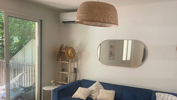 Ett modernt vardagsrum med en blå soffa, ett soffbord i glas, en hänglampa i rotting, en rund spegel och en skjutdörr i glas som leder ut till en balkong.