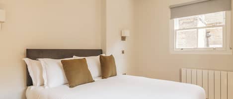 A bedroom with a bed, a nightstand, a window with a view of a brick wall, and a radiator.
