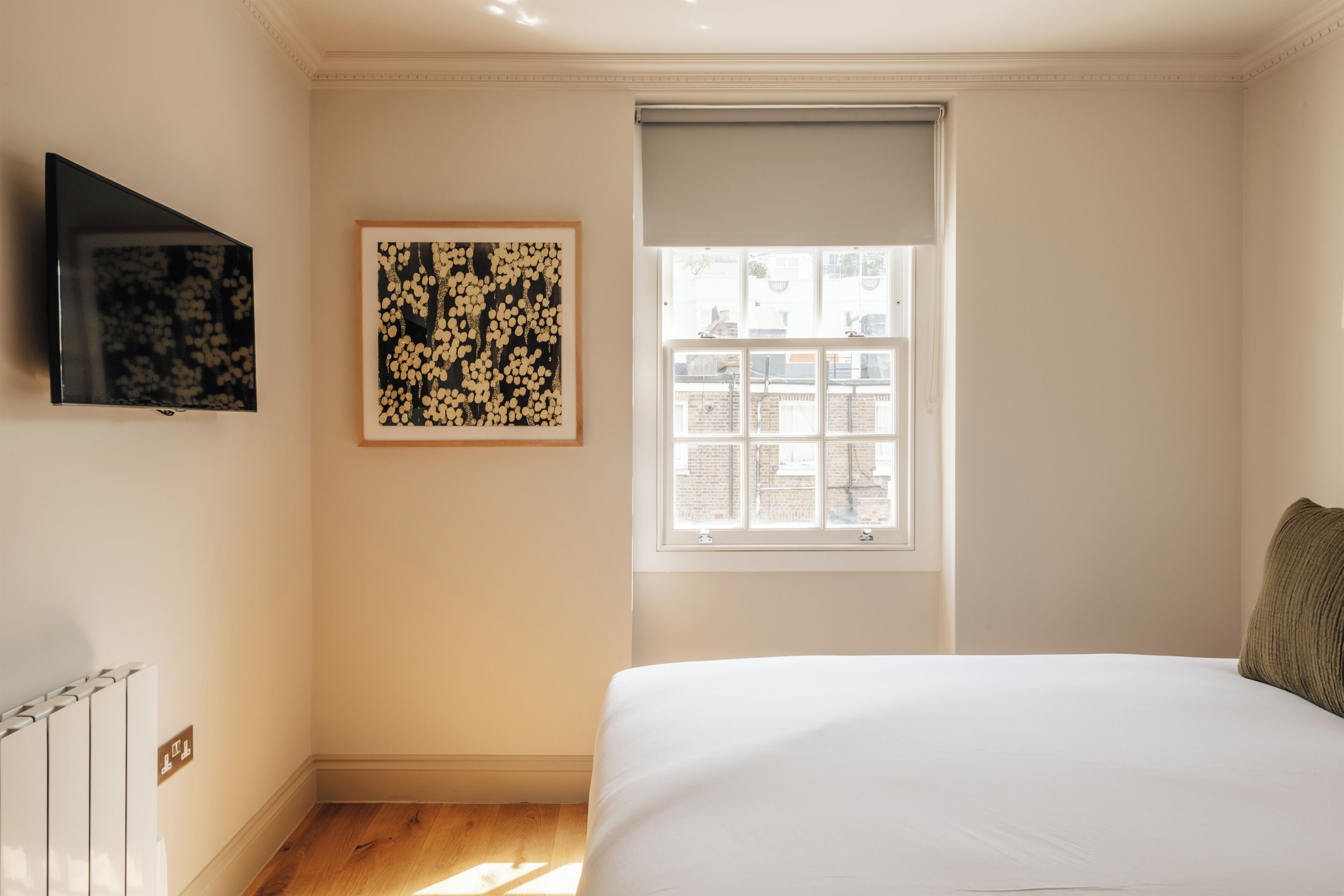 A bedroom with a bed, a television mounted on the wall, a window with a view of buildings, and a framed artwork on the wall.