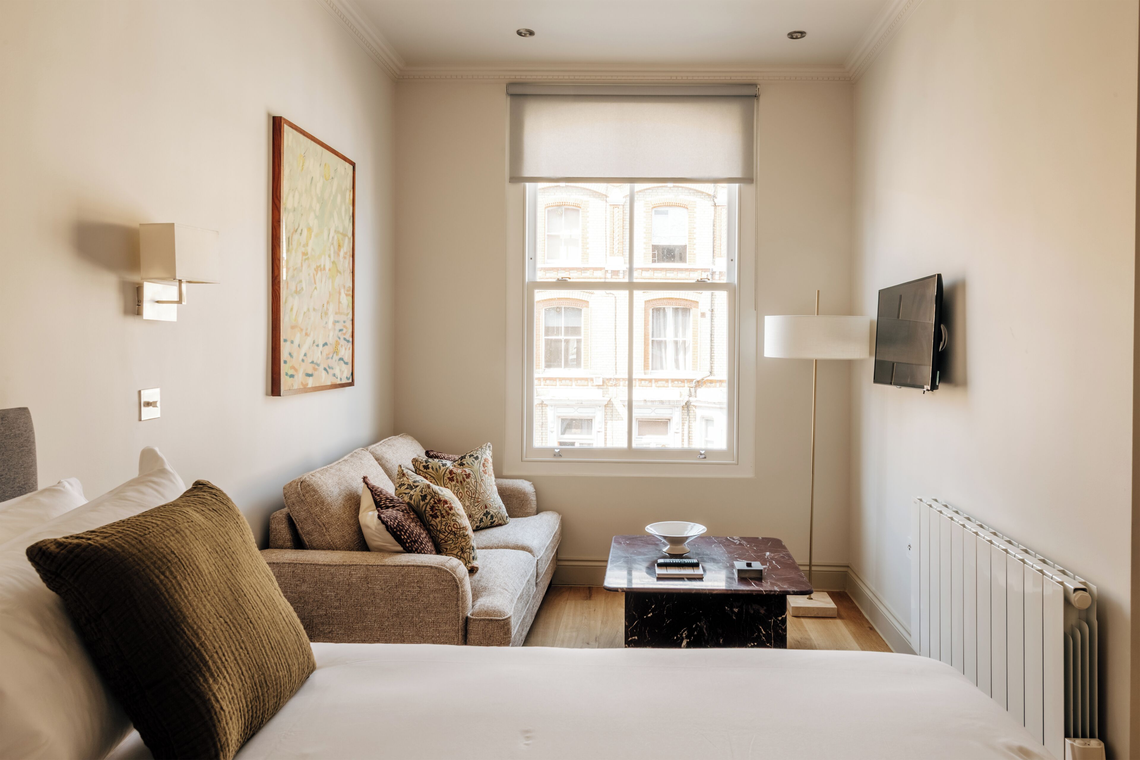 A hotel room with a bed, a sofa, a table with a lamp, a TV, and a window with a view of buildings.