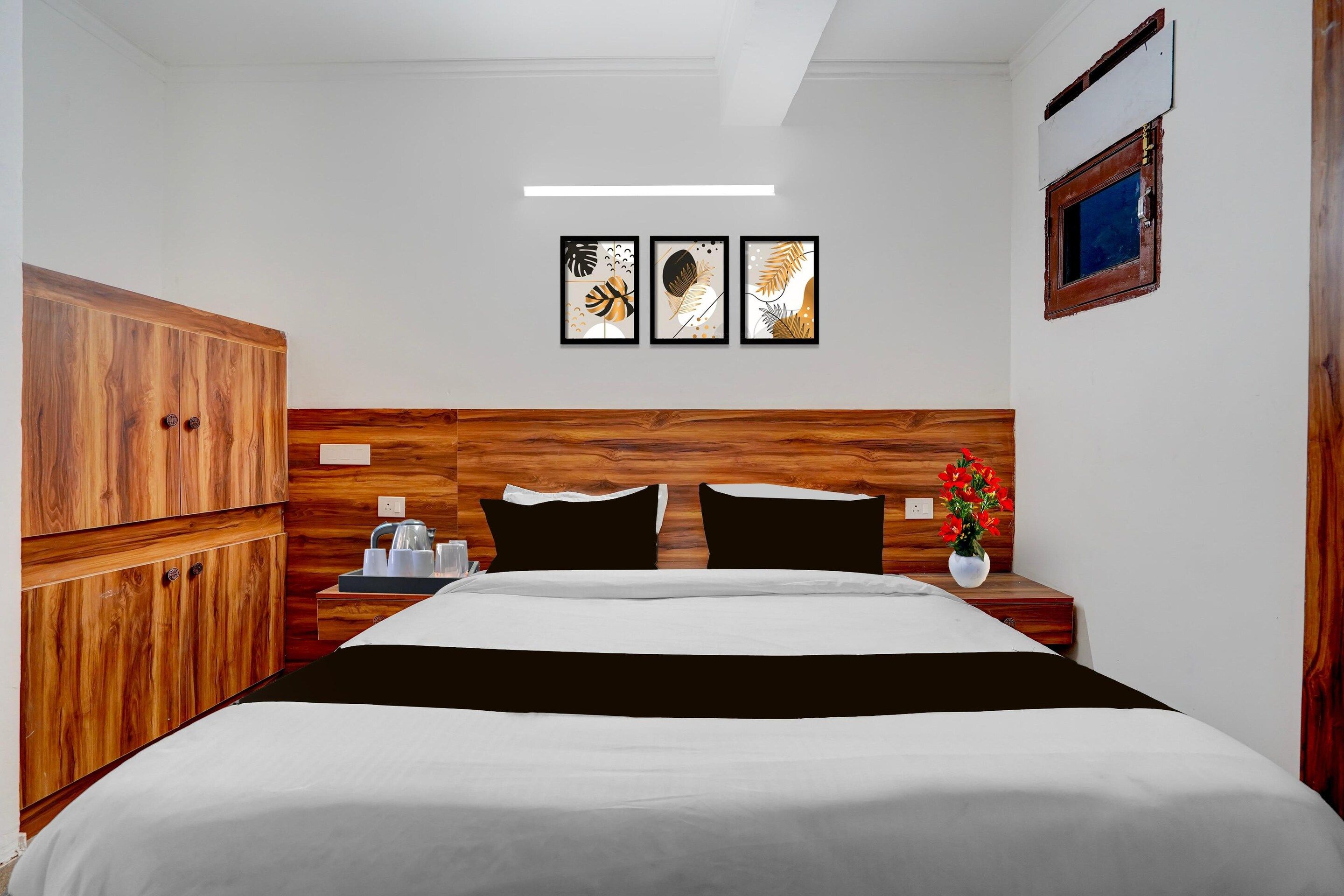 A hotel room with a wooden headboard, a bed with black and white bedding, a nightstand with a kettle, cups, and a vase with flowers.