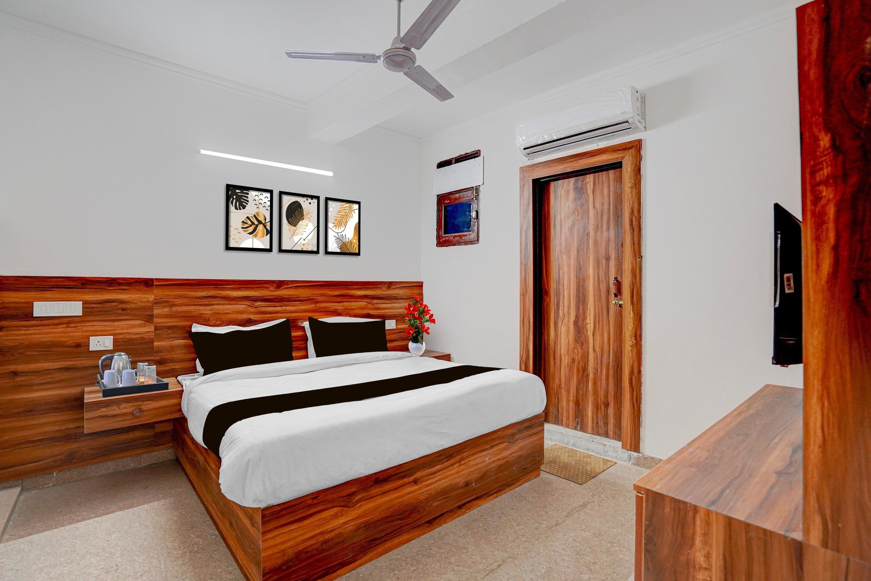 A hotel room with a wooden headboard, a bed with white bedding, a nightstand with a kettle, a wall-mounted air conditioning unit, and a ceiling fan.