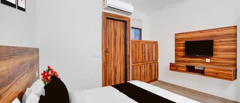 A hotel room with a bed, a wooden wardrobe, a television mounted on a wooden panel, and an air conditioning unit on the wall.