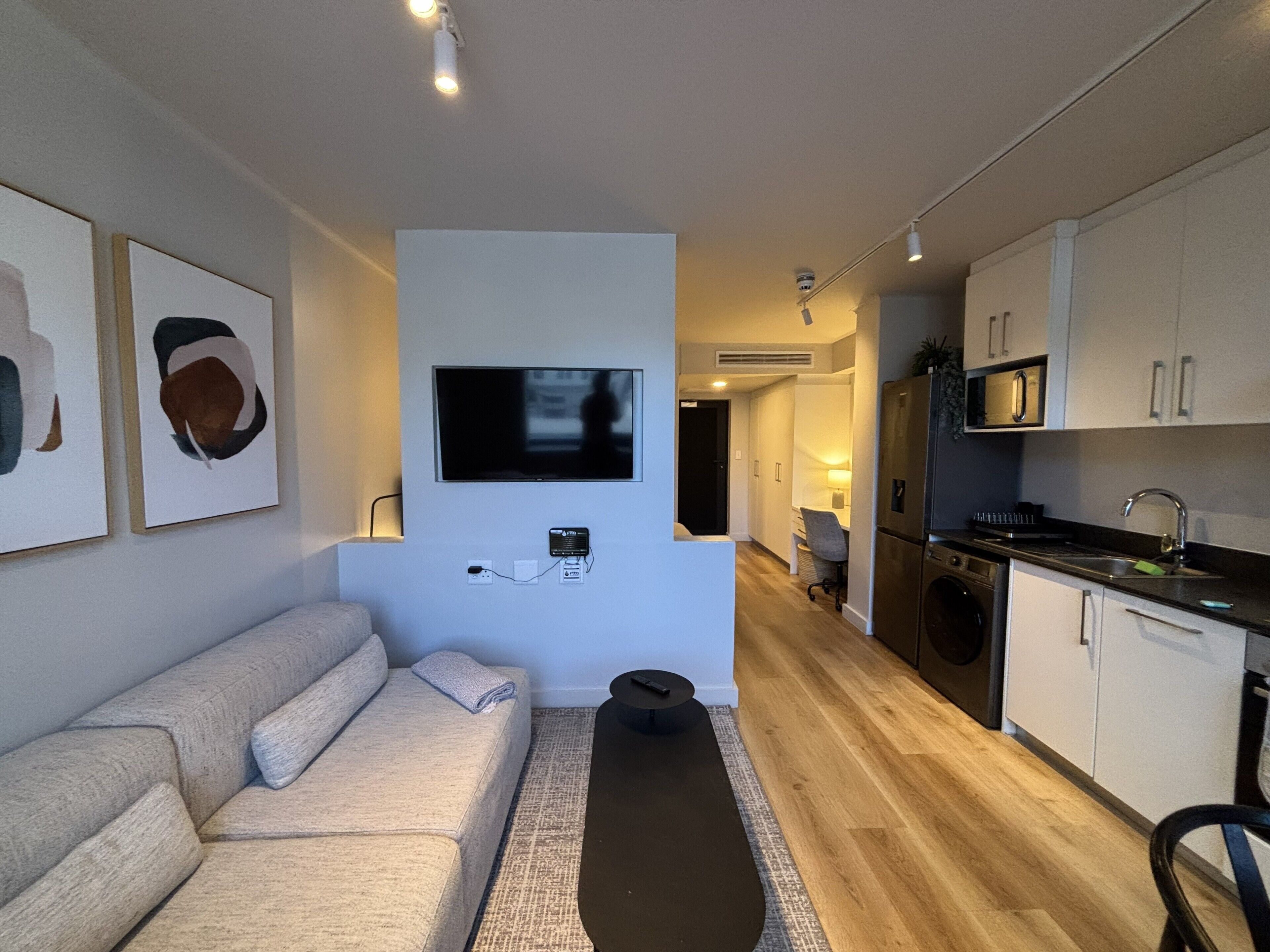 A modern living room with a sofa, a flat-screen TV mounted on the wall, a kitchen area with cabinets and appliances, and a hallway leading to another room.