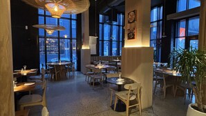 Ein Restaurant mit Tischen, Stühlen, hängenden Lampen und großen Fenstern.