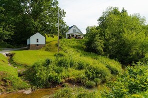 A house with a front porch and a small shed, surrounded by lush greenery and a stream.