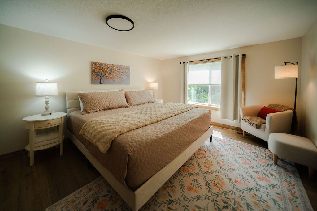 A bedroom with a bed, bedside tables, a chair, a window with curtains, and a painting on the wall.