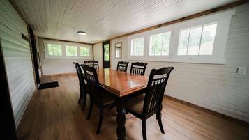 A dining room with a wooden table and chairs, large windows, and a white ceiling.