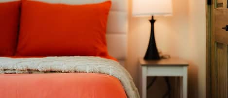 A bed with a white headboard and orange bedding, a bedside lamp, and a small white nightstand.