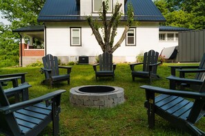 A house with a fire pit surrounded by Adirondack chairs in a grassy yard.