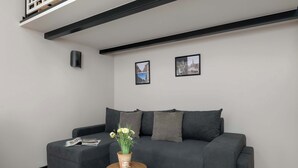Ett modernt vardagsrum med en grå soffa, ett runt sidobord i trä med en vas med blommor och två inramade tavlor på väggen.