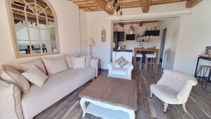 Een gezellige woonkamer met een bank, fauteuil, en salontafel, houten vloeren, en een keuken op de achtergrond.
