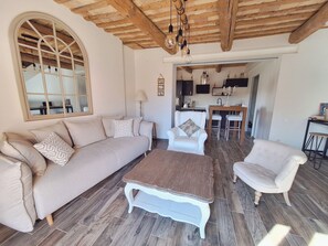 A cozy living room with a sofa, armchair, and coffee table, wooden flooring, and a kitchen in the background. - Provençal house near Uzès with a garden and 2 air-conditioned bedrooms (Le Pin)