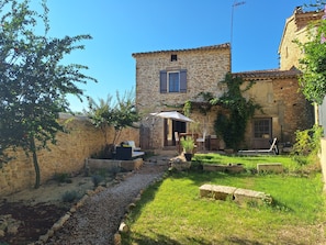 A stone building with a small patio area, a wooden table and chairs, and a garden with a gravel path. - Provençal house near Uzès with a garden and 2 air-conditioned bedrooms (Le Pin)
