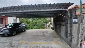 A covered parking area with a car parked under a metal roof, a concrete floor with yellow lines, and a sign indicating 'NOBREGA HOUSE PARKING.'