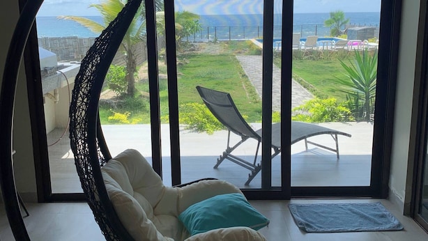A room with a circular hanging chair, a view of a pool and palm trees, and a patio area with a table and chairs.