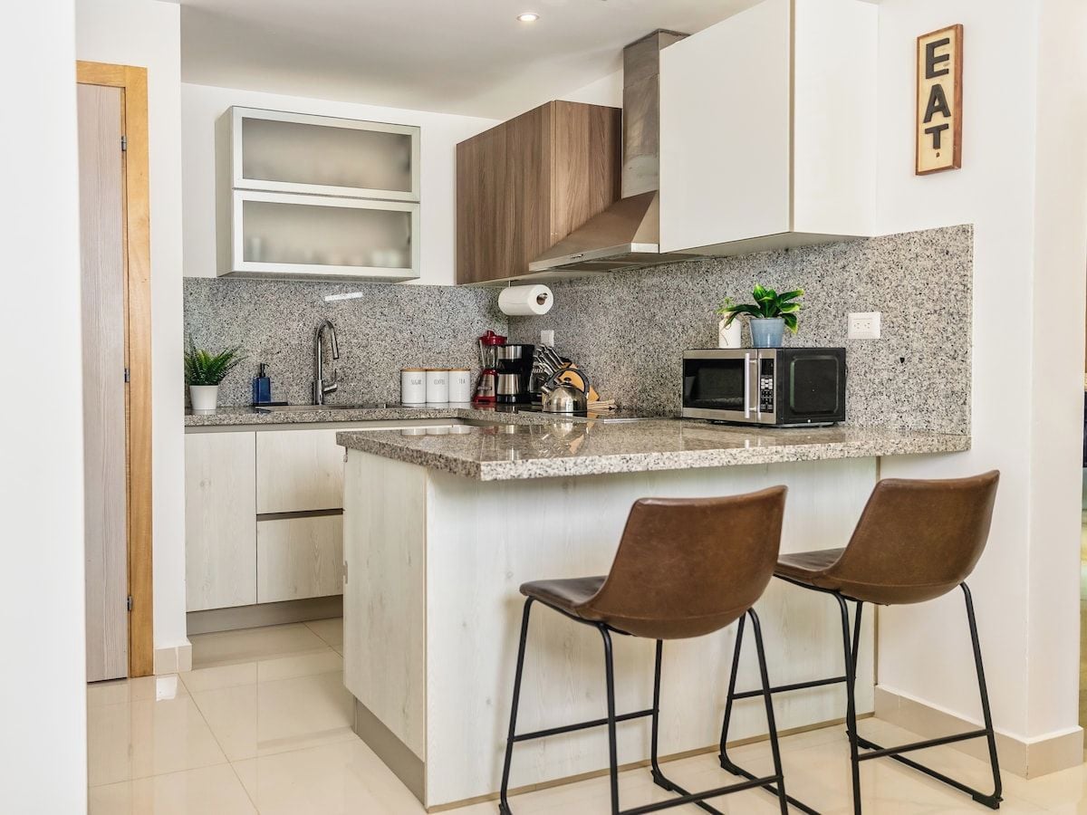 A modern kitchen with a granite countertop, stainless steel appliances, and a bar area with two stools.